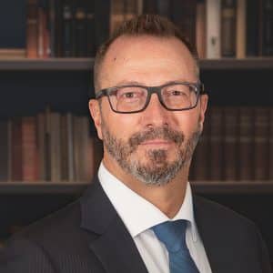 A professional headshot of a man with a beard and glasses, wearing a dark pinstripe suit and blue tie in front of a bookshelf.