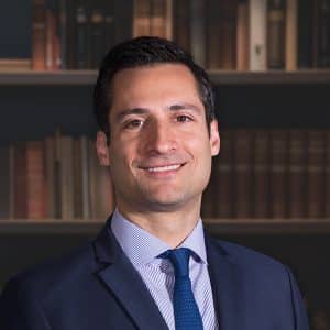 A professional headshot of a smiling man with dark hair, wearing a navy suit, striped shirt, and blue tie in front of a bookshelf.