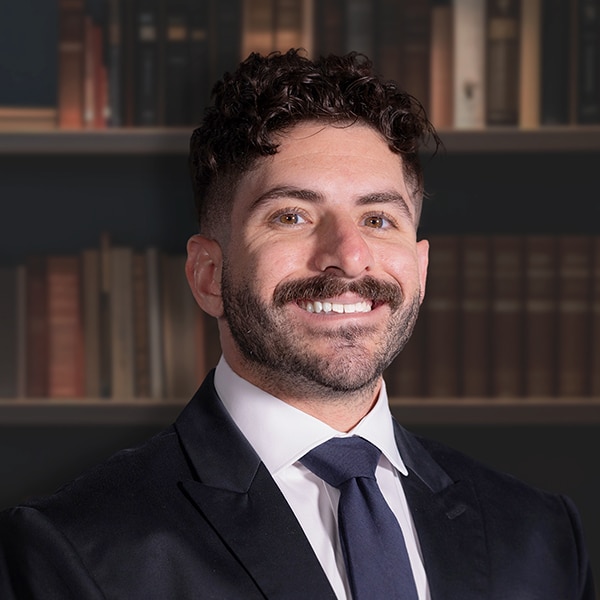 A professional headshot of a smiling man with curly dark hair, a mustache, and a beard, wearing a dark suit and navy tie in front of a bookshelf.