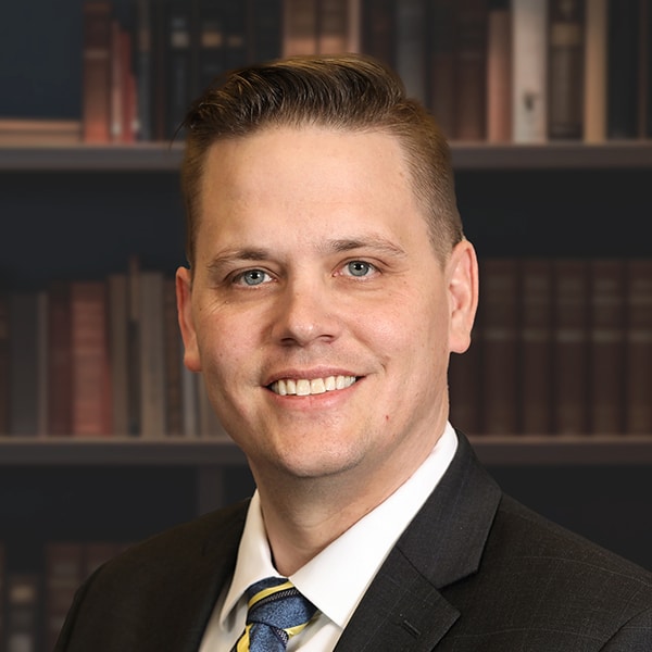 A professional headshot of a smiling man with blue eyes, wearing a dark suit and a blue and yellow striped tie in front of a bookshelf.